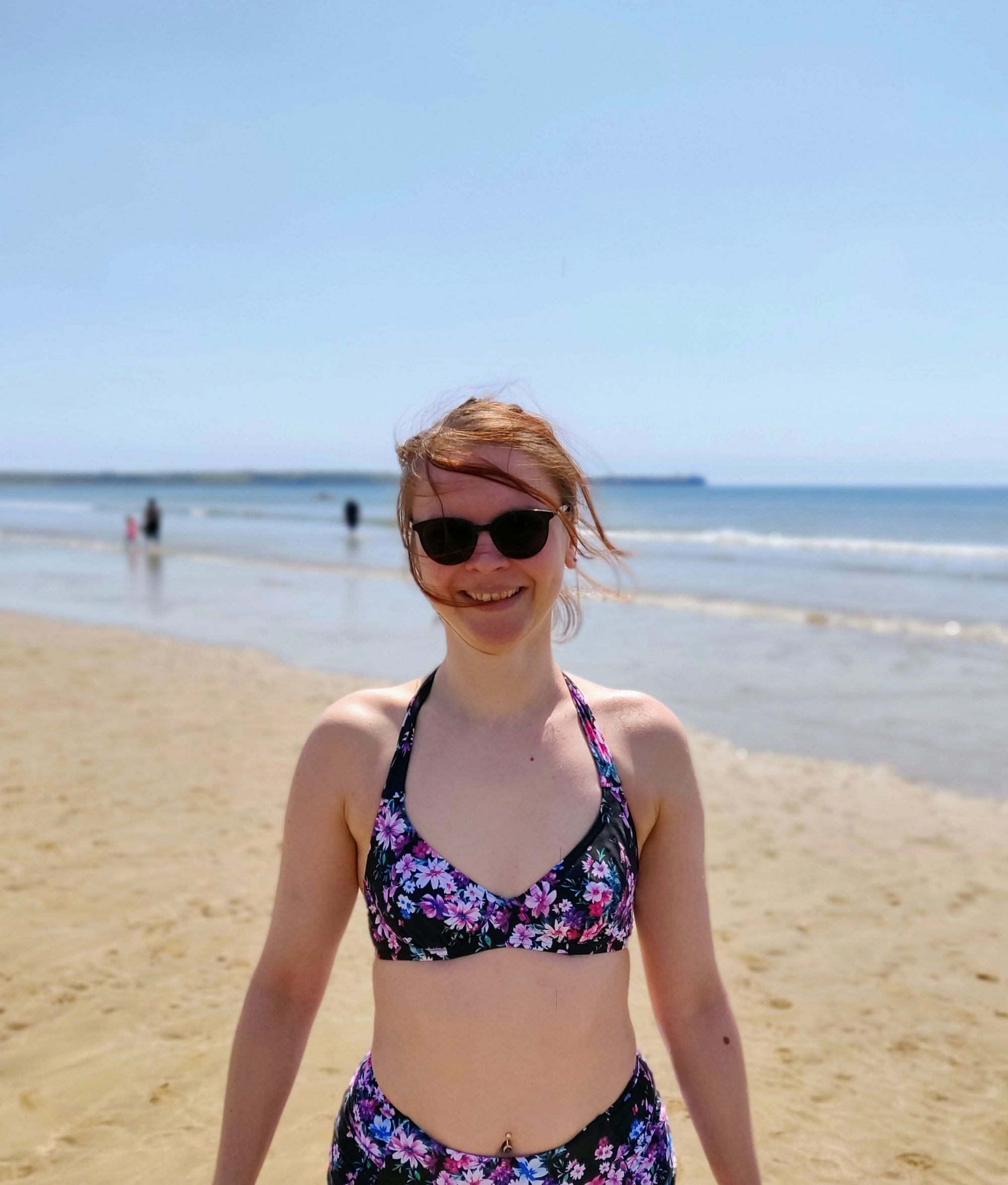 Autorka bloga w okularach przeciwsłonecznych i czarnym bikini w kwiatki stoi na plaży. Za nią widać morze i ludzi, chodzących po nim.